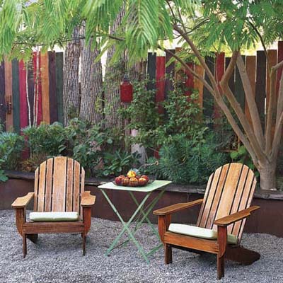 two Adirondack chairs and a small table nestled under the trees in front of a wood fence and plantings