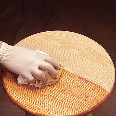 a latex-gloved hand wipes a polyurethane-soaked cloth across a tabletop