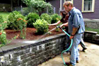 Roger Cook watering newly planted shrubs above a retaining wall with homeowner behind him